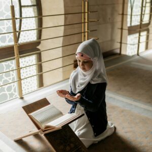 A young Muslim girl in a hijab prays indoors beside an open Quran on a stand.