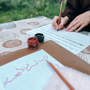 A person practicing calligraphy with ink and pencil on paper in an outdoor setting, focusing on elegant script writing.
